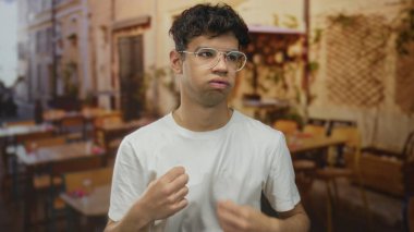 İspanyol gözlüklü genç adam teras restoranında sıcak güneş ışığı altında beyaz gömleği çekiştiriyor; huzursuz..