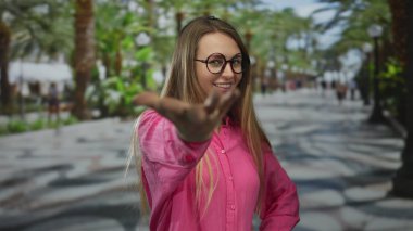 Gözlüklü ve pembe tişörtlü bir kadın canlı park ortamında gülümsüyor, gençlik enerjisi sergiliyor ve özgüvenle işaret ediyor, yemyeşil ve güneşli bir yol ile çerçevelenmiş..