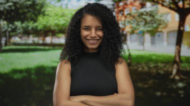 Genç bir kadın güneşli bir park ortamında tuğla bir binanın önünde çıplak omzunu ve çapraz kollarını gösteriyor..