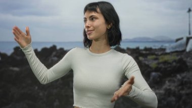 Beyaz ekin giyen kadın, kayalık sahil kenarında uzanmış kolları ve avuç içleri ile yumuşak jestler; davet yansıma dinginliği açıklığı.