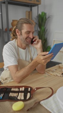 Önlük takan sarışın adam çömlekçilik stüdyosundaki tableti izlerken telefonu kulağına tutuyor..