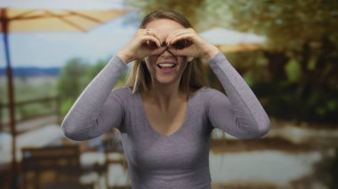 Kadın, güneşli bir kahve restoranının terasında dürbün taklidi yapıyor. Keyif ve rahatlığın somut bir örneği..