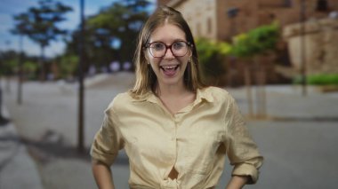 Çıplak kollarıyla dürbün oluşturan bir kadın, güneşli bir şehir sokağında binalarla dolu bir jest yapıyor, neşeyi keşfediyor..