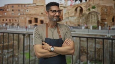 Kameraya gülümseyen antik taş duvarlarla yıkıntılar inşa ederken kolları çapraz kot önlük giyen bir adam..