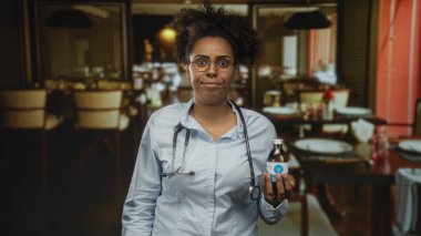 Yemek salonunda steteskoplu ilaç şişesi tutan kadın doktor, gözlük ve nötr ifade; hasta güvenliği endişesi.