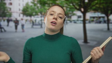 At kuyruklu, gözlüklü, yeşil kazaklı, elinde not defteri olan genç bir kadın..