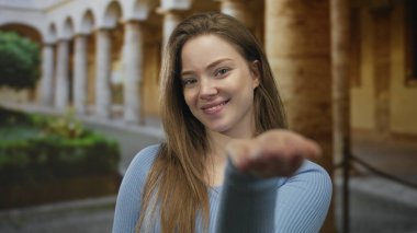 Mavi kazak giyen genç kadın, güneş ışığıyla aydınlanan avluda taş kemer ve kolonlara sahip kameraya doğru elini uzatıyor; dostça bir davet.