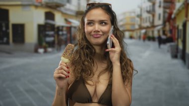 Bikinili İspanyol bir kadın sokakta dikiliyor, akıllı bir telefonla konuşuyor, elinde dondurma tutuyor, gençlik dolu ve canlı bir yaz manzarası yakalıyor..