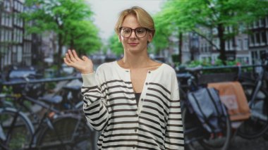 Kadın, park halindeki bisikletlerin ve kanal binalarının yanındaki kalabalık bir caddede, çizgili hırka ve hafif bir gülümsemeyle gözlüğü işaret ediyor; düşünceli sakin bir gülümseme..