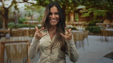 Bir binanın yanındaki bir restoranın terasında iki eliyle kaya boynuzu yapan kadın, neşeli enerji kutlaması özgüveni.