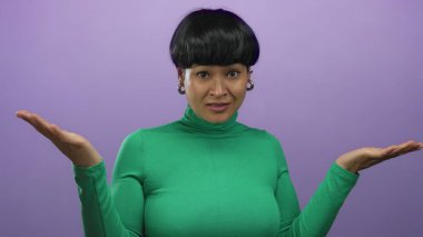 Yeşil elbiseli kadın, izole edilmiş mor arka plana şüpheyle yaklaşıyor. Kısa saçlı ve eşsiz küpeleri var. Minimalist arka planla çelişiyor..