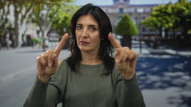 Orta yaşlı İspanyol kadın, güneşli bir şehir sokağında sessiz bir hareketle işaret parmaklarını kaldırıyor..