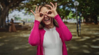 Pembe hırkalı genç sarışın kadın güneşli bir parkta elleri ile dürbün yapıyor, yemyeşil ağaçlar ve neşeli atmosferle çevrili, doğanın tadını çıkartıyor..