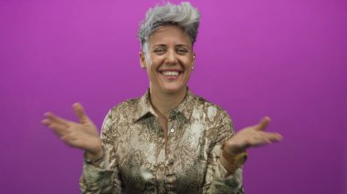 Kısa saçlı, gülümseyen ve iki elini de mor stüdyo ortamında göğsüne koyan beyaz bir kadın..