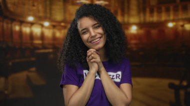 Kapalı bir yerde mor bir gönüllü gömlekle gülümseyen bir kadın sıcacık bir kilise ortamında mutluluk ve neşe ifade ediyor..