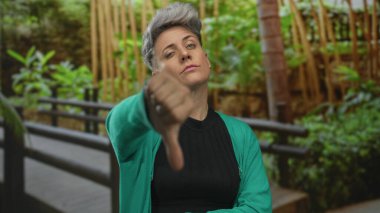 Yeşil hırka giyen kısa saçlı ve uzun kollu bir kadın, yeşil yeşilliklerin ortasında ormanda ahşap bir bankın yanında baş parmak hareketi gösteriyor; tasvip edilmeme.