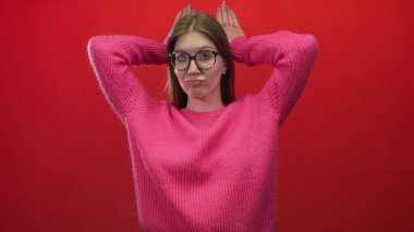 Kırmızı stüdyoda pembe kazaklı, gözlüklü, elleri başının arkasında dinlenen kırmızı gözlü, gevşeme sükuneti olan kadın..