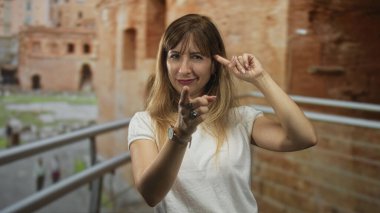 Beyaz tişörtlü genç beyaz kadın kamerayı işaret ediyor ve Roma harabelerinde bir tapınak inşa ediyor..