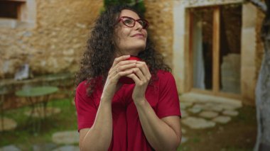 Kıvırcık saçlı, gözlüklü bir kadın, taş bina girişinde kırmızı kupa tutuyor..