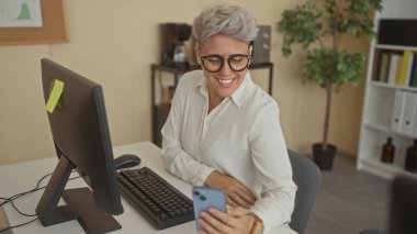 Bir ofis binasında bilgisayar monitörüyle bir masada otururken akıllı telefonu dinleyen bir kadın; bağlantı.