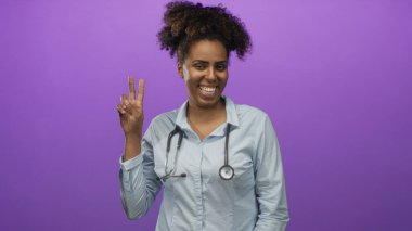 Steteskoplu genç Afrikalı Amerikalı kadın doktor mor stüdyoda barış işareti gösteriyor..