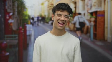 Gözlüklü ve dili açık bir adam renkli kentsel sokak sahnesinde bulanık yayalarla göz kırpıyor; neşeli ruh hali.