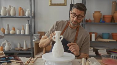 Çömlek atölyesinde elleri ve küçük aletleri olan beyaz bir seramik vazoyu dikkatlice şekillendiren adam parmağıyla kenarı işaret ediyor..