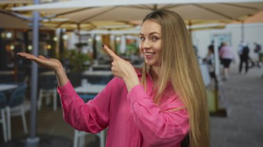 Dışarıdaki bir restoranda pembe bir gömlek giyerken terasta gülümseyen ve işaret eden bir kadın, canlı bir yemek sahnesi ve sıradan bir ortam sergiliyor..