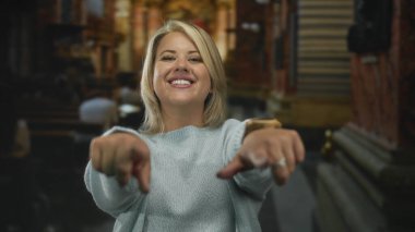 Genç sarışın kadın gülümsüyor ve her iki işaret parmağını dekoratif kilise binasındaki kameraya doğrultuyor;.