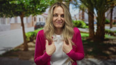 Genç sarışının, açık havada yeşil bir ortamda, şaşırmış bir ifadeyle durduğu parkta kendini işaret eden bir kadın var..