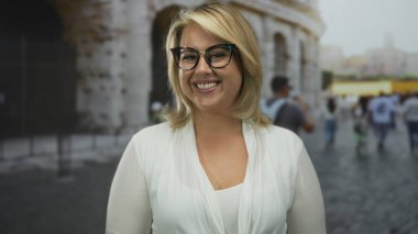 Gözlüklü sarışın kadın gündüz vakti kolezyum binasının dışındaki kamerayı işaret ediyor; özgüveni vurgula.