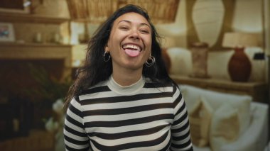 Dilini çıkaran ve stüdyoda parmaklarını yanaklarına gösteren bir kadın. Ampuller ve kanepe görünür..