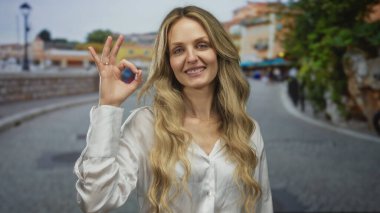Açık havada gülümseyen sarışın bir kadın sokakta jestler yapıyor beyaz bir gömlek giyerken işaret veriyor, mutluluğu ve kendine güveni etkileyici bir ifadeyle somutlaştırıyor..