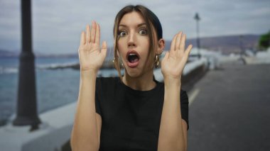 Genç İspanyol kadın gün batımında deniz kenarında bir gezinti alanında avuç içi ile gözlerini kapar; sürpriz merak.