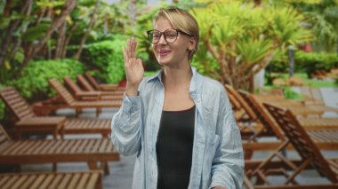 Kadın yüzün yanında el sallıyor, tahta şezlonglu gözlük takıyor ve binadaki havuz başında tropikal bitkiler takıyor..