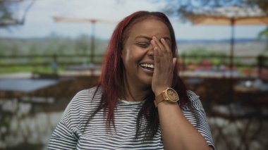 Afro-Amerikalı kadın, açık hava restoranlarının terasında kahkahalarla gülen ağzını kapatıyor..