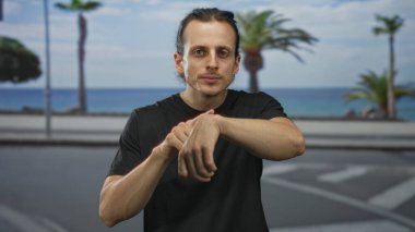 Siyah tişörtlü, uzun saçlı, okyanus ve palmiye ağaçlarının yanındaki sokakta parmak bileğe gezen genç adam sabırsızlık kontrolü yapıyor..
