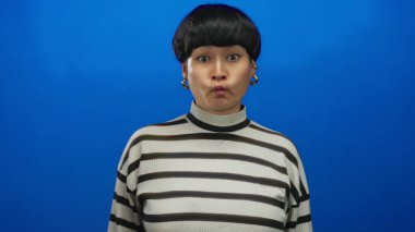 Mavi arka planda, çizgili süveter ve küpe takan, renkli ve izole bir ortamda sürpriz yapan bir kadın..