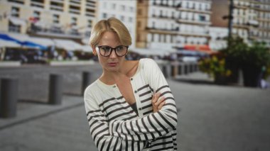 Kolları çapraz olan ve kaldırım taşları ve apartman binalarının yanındaki kaldırım taşından bir sokakta duran gözlüklü kadın, şüpheli bir bakışla kameraya bakıyor; şüphecilik..