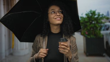Sokakta yağmurlu bir günde elinde şemsiye ve kahve bardaklarıyla gülümseyen bir kadın..