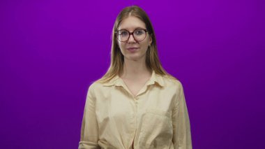 Sarı saçlı, bej gömlekli ve gözlüklü bir kadın mor bir stüdyoda hafif bir gülümsemeyle kollarını kavuşturuyor..