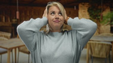 Sarı saçlı, açık renk kazaklı bir kadın bina terasında kulaklarını kapatmak için ellerini kaldırıyor..