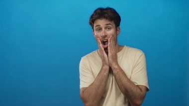 Elleri mavi stüdyoda ağzını kapatan adam, bej tişört, görünür kıllar ve ağzı açık bir şekilde şok hareketi gösteren adam; sürpriz..