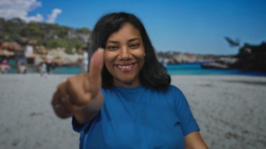 Mavi gömlekli ve gülümseyen bir kadın kumlu sahilde olumlu bir jest yapıyor..