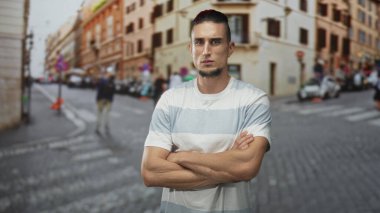 Şehir merkezinde kaldırım taşı caddesinde kolları çaprazlanmış bir adam, çizgili gömlek ve keçi sakalı görünür; özgüven dayanıklılığı.