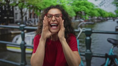 Kanal köprüsünde elleri ağızdan ağıza kırmızı gömlek ve gözlük takan bir kadın. İletişim heyecanı..