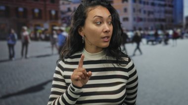 Kadın, meşgul bir şehir caddesinde işaret parmağını kaldırırken kaşlarını kaldırıyor ve yan yan bakıyor; dikkat.
