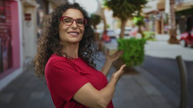 Kıvırcık saçlı ve kırmızı gözlüklü genç İspanyol kadın kalabalık bir sokak sahnesinde kollarını açıyor..