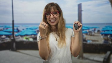 Kadın arabanın anahtarını sıkılmış yumruğuyla tutuyor ve kalabalık bir sahil yürüyüşünde mavi şemsiyeler ve denizle gülümsüyor..