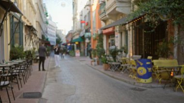 Bulanık sokak kafe sahnesi sığ bokeh, odaklanmış vitrin önleri, açık alan oturma ve kaldırım taşı yolu; arkaplan sakin telif alanı.